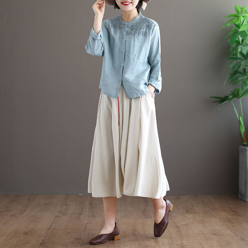 Woman wearing a light blue shirt and beige skirt standing against a gray wall with a plant to the right.