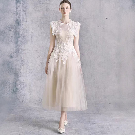 A woman models a sleeveless champagne tulle dress with intricate 3D floral lace appliques on the bodice and waist, set against a textured gray studio backdrop.