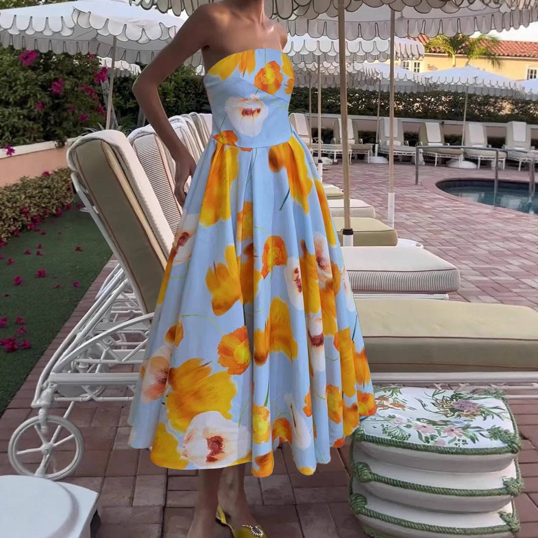 A woman wearing a strapless light blue A-line dress with a large yellow and white floral print, standing in a resort poolside setting with lounge chairs.